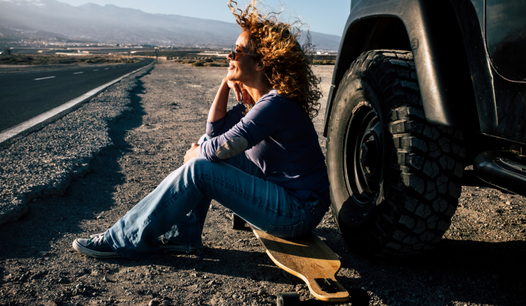 Adult woman witting on a longboard outside the car with long road in background